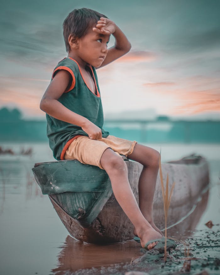 A young boy sits on a wooden boat by a misty riverside in Nepal, gazing into the distance.