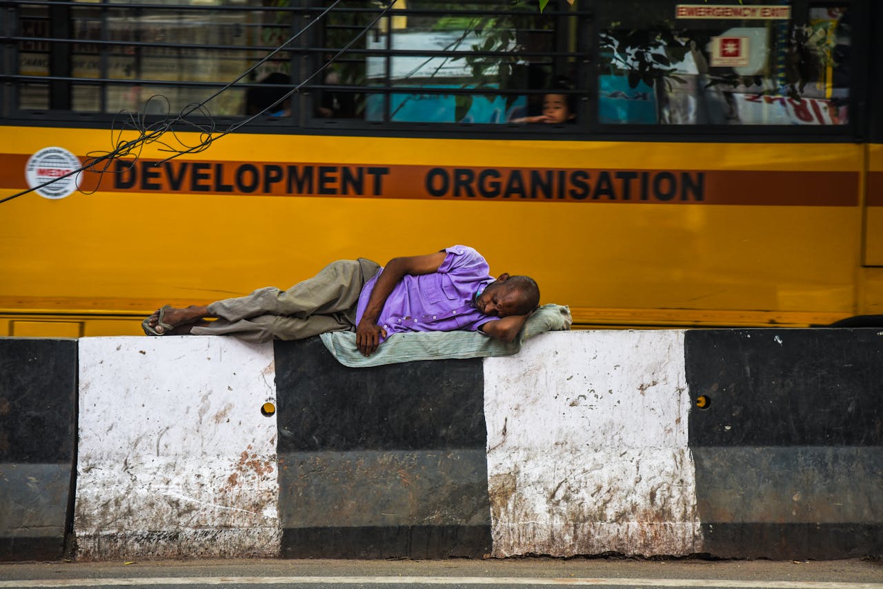 services-02 Elderly man sleeping on a concrete barricade beside a moving yellow bus in an urban setting.